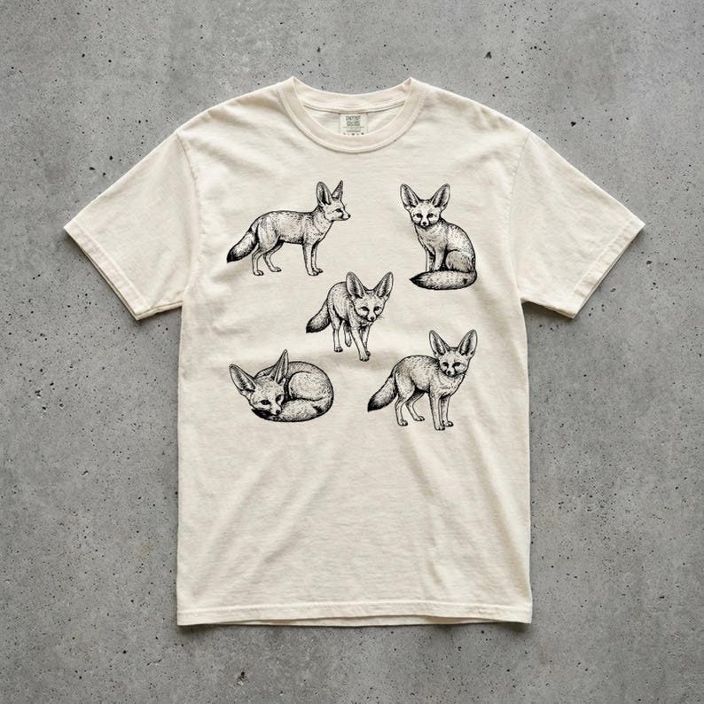 Ivory graphic t-shirt featuring a vintage engraved style illustration of multiple fennec foxes arranged in different poses. The foxes are shown sitting, walking, and curled up, with detailed linework creating a naturalist wildlife illustration and desert animal aesthetic. Laid flat on a concrete surface.