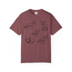 Brick red graphic t-shirt featuring a vintage engraved style illustration of multiple fennec foxes arranged in different poses. The foxes are shown sitting, walking, and curled up, with detailed linework creating a naturalist wildlife illustration and desert animal aesthetic.