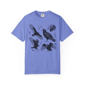 Flo blue Comfort Colors t-shirt with a vintage engraved illustration of five vultures including one soaring with wings outstretched, one standing with wings half-spread, a close-up portrait showing bare head and hooked beak, one landing with wings fanned, and one spreading wings low in a dominance display, surrounded by bare winter branches and scattered feathers in an antique wildlife field guide etching style.