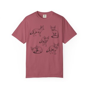 Crimson red graphic t-shirt featuring a vintage engraved style illustration of multiple fennec foxes arranged in different poses. The foxes are shown sitting, walking, and curled up, with detailed linework creating a naturalist wildlife illustration and desert animal aesthetic.