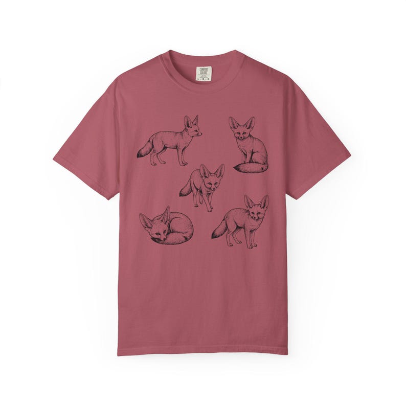 Crimson red graphic t-shirt featuring a vintage engraved style illustration of multiple fennec foxes arranged in different poses. The foxes are shown sitting, walking, and curled up, with detailed linework creating a naturalist wildlife illustration and desert animal aesthetic.
