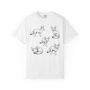 White graphic t-shirt featuring a vintage engraved style illustration of multiple fennec foxes arranged in different poses. The foxes are shown sitting, walking, and curled up, with detailed linework creating a naturalist wildlife illustration and desert animal aesthetic.