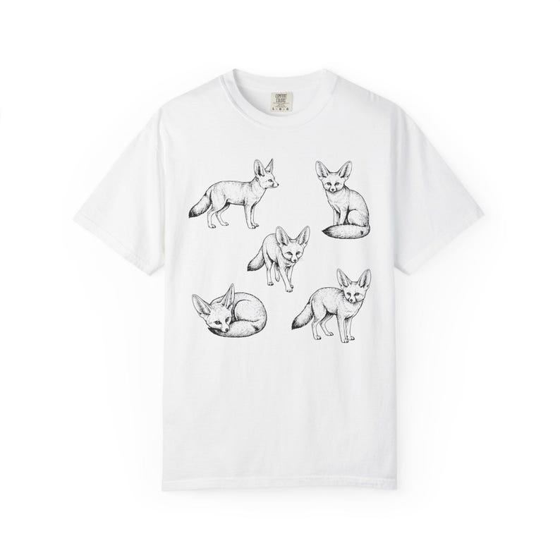 White graphic t-shirt featuring a vintage engraved style illustration of multiple fennec foxes arranged in different poses. The foxes are shown sitting, walking, and curled up, with detailed linework creating a naturalist wildlife illustration and desert animal aesthetic.