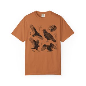 Yam orange Comfort Colors t-shirt with a vintage engraved illustration of five vultures including one soaring with wings outstretched, one standing with wings half-spread, a close-up portrait showing bare head and hooked beak, one landing with wings fanned, and one spreading wings low in a dominance display, surrounded by bare winter branches and scattered feathers in an antique wildlife field guide etching style.