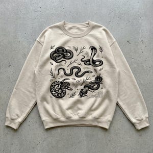 May include: A beige crew neck sweatshirt with a black and white illustration of several snakes in different poses, accompanied by sprigs and leaves. The snakes are depicted in a detailed, artistic style, creating a striking contrast against the neutral background.