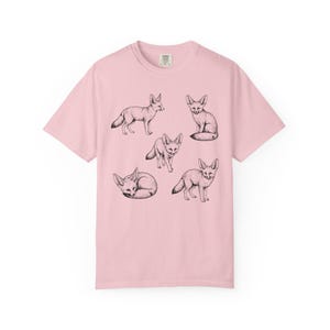 Light pink graphic t-shirt featuring a vintage engraved style illustration of multiple fennec foxes arranged in different poses. The foxes are shown sitting, walking, and curled up, with detailed linework creating a naturalist wildlife illustration and desert animal aesthetic.