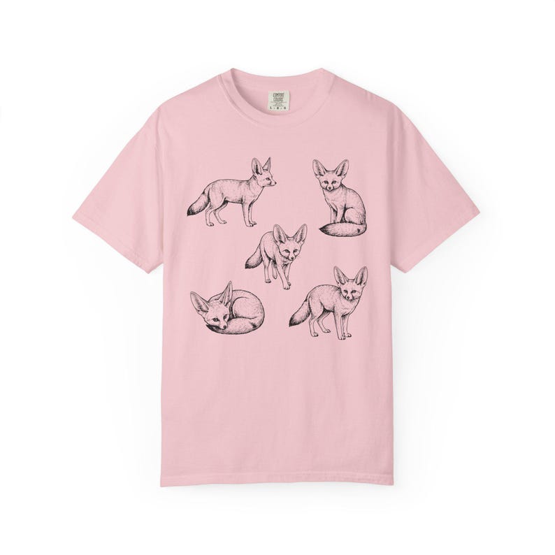 Light pink graphic t-shirt featuring a vintage engraved style illustration of multiple fennec foxes arranged in different poses. The foxes are shown sitting, walking, and curled up, with detailed linework creating a naturalist wildlife illustration and desert animal aesthetic.