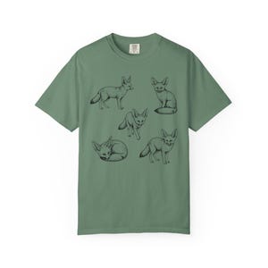 Light green graphic t-shirt featuring a vintage engraved style illustration of multiple fennec foxes arranged in different poses. The foxes are shown sitting, walking, and curled up, with detailed linework creating a naturalist wildlife illustration and desert animal aesthetic.