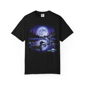 Black Comfort Colors garment-dyed t-shirt featuring a full-color 90s airbrush-style circular vignette illustration of a common loon swimming on a moonlit violet northern lake with a second loon in flight, blue water lilies, pine silhouettes, and a full moon — unisex graphic tee.