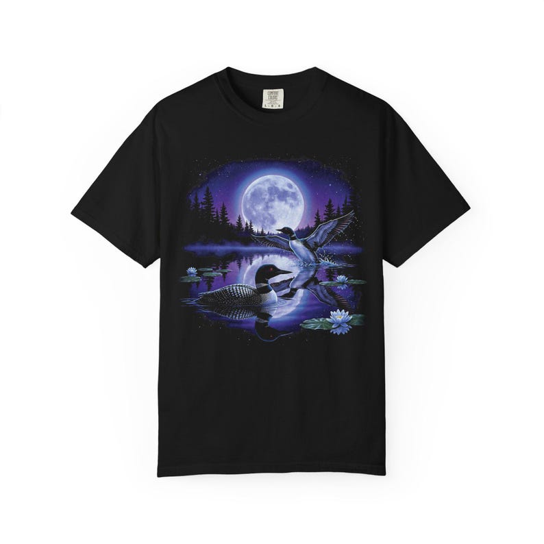 Black Comfort Colors garment-dyed t-shirt featuring a full-color 90s airbrush-style circular vignette illustration of a common loon swimming on a moonlit violet northern lake with a second loon in flight, blue water lilies, pine silhouettes, and a full moon — unisex graphic tee.