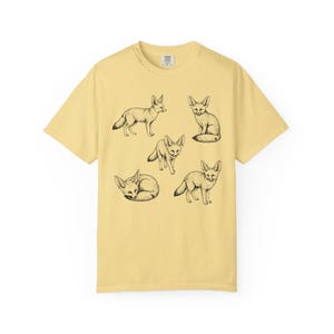 Butter yellow graphic t-shirt featuring a vintage engraved style illustration of multiple fennec foxes arranged in different poses. The foxes are shown sitting, walking, and curled up, with detailed linework creating a naturalist wildlife illustration and desert animal aesthetic.