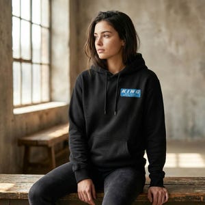 Könnte beinhalten: Ein schwarzer Kapuzenpullover mit einer Vordertasche und einer Kapuze. Der Pullover hat einen blauen rechteckigen Aufnäher auf der Brust mit dem Wort "KING" in weißen Buchstaben. Die Person trägt eine dunkle Jeans und sitzt auf einer Holzbank.