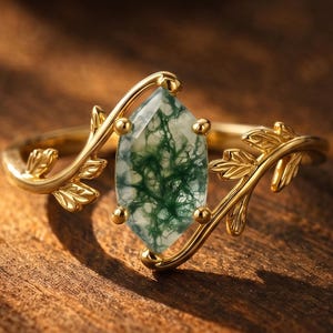 May include: A gold ring with a central, elongated, green and white moss agate stone. The ring features a delicate, leaf-like design on the band, with the stone set in a gold bezel. The ring is displayed on a wooden surface.