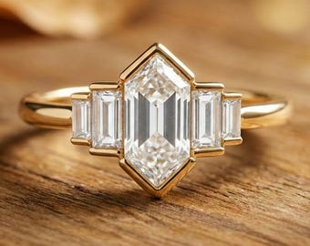 May include: A gold ring featuring a large, clear, emerald-cut center stone, flanked by smaller rectangular stones. The ring has a geometric design and is set against a wooden surface.