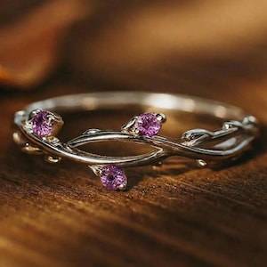 May include: A silver ring with a delicate vine design, adorned with three small, round, purple gemstones. The ring's band is thin and polished, and the gemstones are set in the vine's leaves. The ring is displayed on a wooden surface.