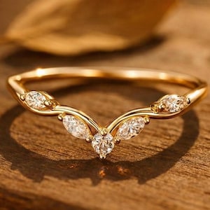Puede incluir: Un anillo dorado con un diseño en forma de V. El anillo presenta varias piedras preciosas claras y brillantes. Las piedras están engastadas en un patrón elegante. El anillo se muestra sobre una superficie de madera.