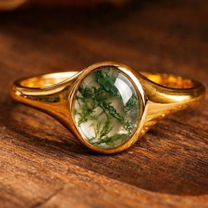 Puede incluir: Un anillo de oro con una piedra de ágata musgosa de forma ovalada. La piedra presenta inclusiones verdes y blancas, engastada en una banda de oro pulido. El anillo se muestra sobre una superficie de madera, resaltando su diseño y elementos naturales.