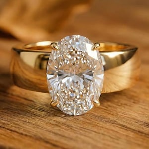 Puede incluir: Un anillo de oro con un gran diamante de talla ovalada. El diamante es el punto focal, engastado en un engaste clásico de cuatro puntas. La banda es ancha y pulida, reflejando la luz. El anillo está sobre una superficie de madera.