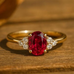 Puede incluir: Un anillo de oro con un gran rubí de talla ovalada como pieza central. El rubí es de color rojo intenso y está flanqueado por piedras preciosas más pequeñas, redondas y transparentes a cada lado. El anillo está engastado en una banda de oro.