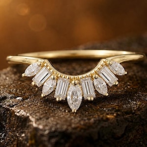 May include: A gold ring featuring a curved design, adorned with multiple clear gemstones. The gemstones are a mix of shapes, including marquise and baguette cuts, arranged in a fan-like pattern. The ring is displayed on a dark, textured surface.