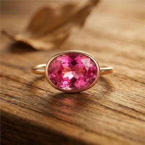 Puede incluir: Un anillo de oro con una gran piedra preciosa rosa de talla ovalada. La piedra preciosa está engastada en un bisel y tiene múltiples facetas que reflejan la luz. El anillo se muestra sobre una superficie de madera.