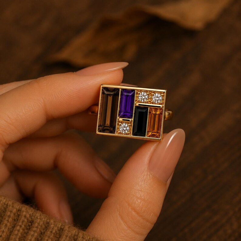 Puede incluir: Un anillo de oro con una cara rectangular con un dise&ntilde;o geom&eacute;trico. El dise&ntilde;o incorpora piedras rectangulares en tonos marr&oacute;n, morado, negro y naranja, junto con dos grupos de peque&ntilde;as piedras blancas. El anillo es sostenido por una persona.