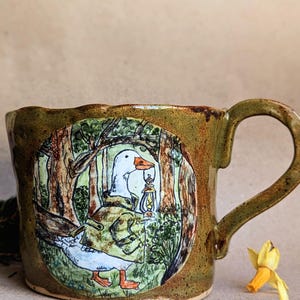 May include: A ceramic mug with a whimsical illustration of a goose in a forest scene, holding a lantern. The mug has a brown and green color scheme with a wavy rim and a large handle. The mug is approximately 4 inches tall.