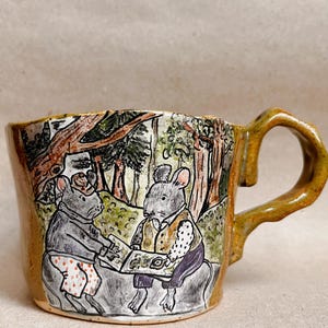 May include: A ceramic mug with a glossy mustard yellow glaze and a hand-drawn illustration of two anthropomorphic mice sitting in a forest setting. One mouse wears polka-dot pants and the other wears a vest, both looking at an open book.