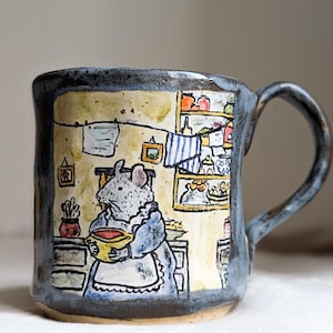 May include: Hand-painted ceramic mug featuring a mouse in a kitchen scene. The mug has a blue and grey exterior and a detailed interior illustration. The mouse is holding a bowl. A unique and charming kitchenware item, perfect for tea or coffee.
