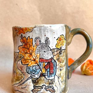 Könnte beinhalten: Ein Keramikbecher mit einer handgemalten Illustration einer grauen Maus, die einen roten Schal und eine blaue Jacke trägt und ein großes orangefarbenes Herbstblatt hält. Der Hintergrund zeigt eine Landschaft mit einem Baum in Erdtönen.
