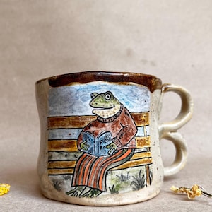 Könnte beinhalten: Ein gesprenkelter Keramikbecher mit doppeltem Henkel, der eine handgemalte Illustration eines Frosches in einem braunen Pullover und einer gestreiften Hose zeigt, der auf einer Bank sitzt und ein blaues Buch mit dem Wort Frog auf dem Einband liest.