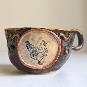 Può includere: Ciotola da zuppa in ceramica fatta a mano con esterno marrone e interno color crema. La ciotola presenta un'immagine dipinta a mano di un gallo all'interno di una cornice ovale. Il bordo ha un design decorativo giallo e marrone e un manico.