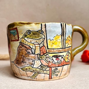 Pode incluir: Uma caneca de cerâmica com uma ilustração pintada à mão de um sapo verde vestindo uma jaqueta marrom e um cachecol listrado. O sapo está sentado a uma mesa com uma xícara de chá e um bule perto de uma janela. A caneca tem um acabamento brilhante.