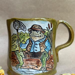 May include: A ceramic mug with a speckled olive green glaze featuring a hand-painted illustration of a frog wearing a hat, blue shirt, and brown vest. The frog holds a net while sitting on a log near cattails by a blue water surface.
