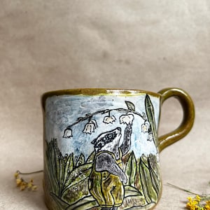 May include: A ceramic mug with a glossy olive green handle and rim, featuring a hand-painted illustration of a badger wearing yellow overalls standing in a field of green leaves and reaching up toward white lily of the valley flowers.