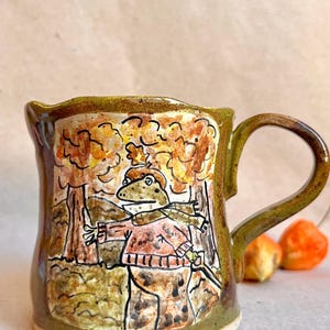Peut inclure: Un mug en céramique avec une glaçure vert olive brillante et une illustration peinte à la main d'une grenouille portant un pull et une écharpe dans une forêt automnale. La scène montre des arbres orange et jaunes, avec une anse courbée.