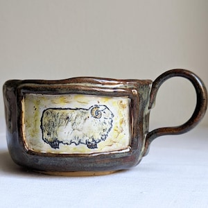 Puede incluir: Taza de cerámica hecha a mano con exterior marrón y un panel rectangular con una ilustración de oveja. La taza tiene un asa redondeada y un diseño rústico único. La oveja está representada en blanco y negro sobre un fondo amarillo.