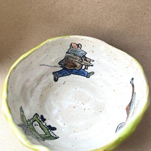 Könnte beinhalten: Eine handgefertigte Keramikschale mit gesprenkeltem weißem Innenraum, verziert mit handgemalten Illustrationen einer Maus in brauner Jacke und blauer Hose, einem grünen Frosch und einem kleinen Vogel. Die Schale hat einen welligen Rand und eine lindgrüne Außenseite.