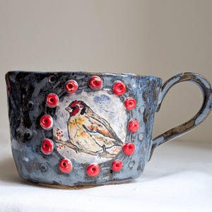 Peut inclure: Tasse en céramique faite à la main avec une illustration d'oiseau. La tasse est principalement bleue avec une surface texturée et une anse. L'oiseau est représenté au centre, entouré d'accents rouges. Une pièce unique pour savourer des boissons.