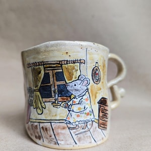 Può includere: Una tazza in ceramica con una stravagante illustrazione di un topo che tiene in mano una candela vicino a una finestra. La tazza presenta una base color crema con un manico marrone e un disegno dettagliato di una scena interna accogliente.