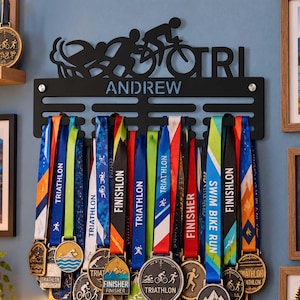 May include: A black metal wall-mounted medal hanger featuring silhouettes of a swimmer, cyclist, and runner above the text TRI and the name ANDREW. The hanger displays numerous colorful ribbons with various triathlon finisher medals.