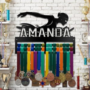 May include: Black metal medal display with the name "AMANDA" and a swimmer silhouette. The display has colorful ribbons holding various medals. The background is a white brick wall, with trophies on shelves.