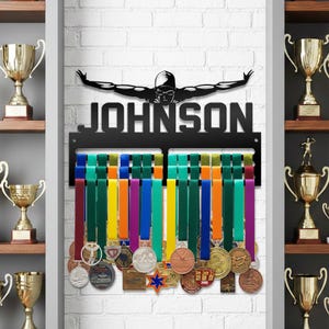 Könnte beinhalten: Eine schwarze Metall-Medaillen-Anzeige mit dem Namen "JOHNSON" und einer Schwimmer-Silhouette. Die Anzeige verfügt über farbige Bänder, die verschiedene Medaillen halten. Trophäen sind auf Regalen im Hintergrund zu sehen.