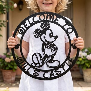 Op de afbeelding: Een rond zwart metalen bord met een uitgesneden ontwerp van Mickey Mouse in het midden. De tekst "WELCOME TO" staat in een boog aan de bovenkant en "OILL'S CASTLE" aan de onderkant. Kleine Mickey Mouse-hoofdsilhouetten staan aan de zijkanten.