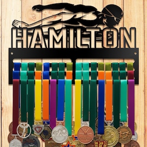 Könnte beinhalten: Schwarzes Metall-Medaillen-Display mit dem Namen "HAMILTON" und einer Schwimmer-Silhouette. Das Display hat mehrere farbige Bänder zum Aufhängen von Medaillen. Verschiedene Medaillen mit unterschiedlichen Designs sind sichtbar, die an den Bändern hängen.