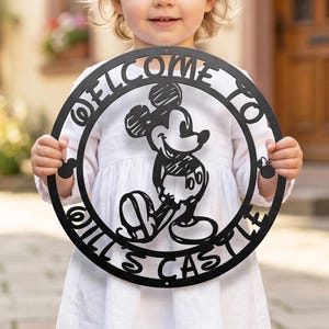Könnte beinhalten: Ein kreisförmiges schwarzes Metallschild mit einem Scherenschnitt einer Zeichentrick-Mausfigur in der Mitte. Der Text "WELCOME TO" ist oben und "OILL'S CASTLE" unten bogenförmig angeordnet. Kleine Mausohren-Silhouetten befinden sich an den Seiten.