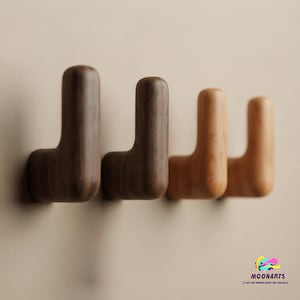 Könnte beinhalten: Vier L-förmige Wandhaken aus Holz, die in einer horizontalen Reihe angeordnet sind. Die beiden linken Haken sind dunkelbraun mit sichtbarer Holzmaserung, während die beiden rechten Haken einen helleren, goldbraunen Ton haben. Der Text MOONARTS ist sichtbar.