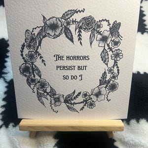 以下が含まれることがあります： 黒い花のリースデザインと「The horrors persist but so do I」のテキストが特徴の、正方形のオフホワイトカード。カードは小さな木製のイーゼルに飾られ、白黒のチェック柄の背景に配置されています。
