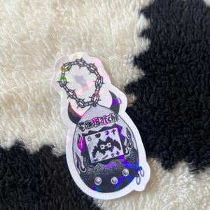 May include: A holographic sticker featuring a stylized Tamagotchi design. The sticker has a black and white color scheme with a chain and horns. The text "Tamabatchi" is visible on the top of the sticker.