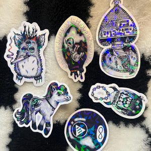 May include: A collection of holographic stickers with various designs. The stickers include a three-eyed creature smoking a cigarette, an alien in an egg, a house, a pony, a magic 8-ball, and a potion bottle.